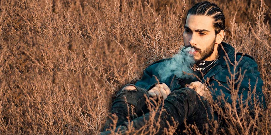 a man with braided hair vaping outdoors and sitting on a grass field