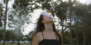 a woman wearing a black top vaping and exhaling smoke outdoors with trees in the background